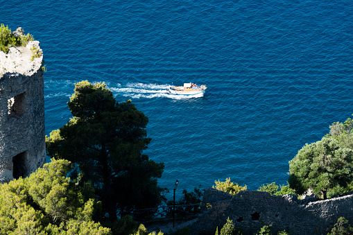 View from above, stunning view of a gozzo boat with tourists on board sailing on a blue water. Defocused Torre dello Ziro in the foreground. The 