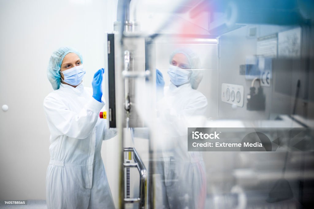 Pharmaceutical industry and drug manufacturing A woman wearing proper safety clothes seen taping on the screen of a big complex machine in pharmaceutical industry while her reflection is visible on the machine. Medicine Stock Photo