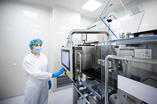 Female employee  in pharmaceutical industry seen standing in front of the screen on the very complex machine and pushing a button while wearing protective white suit, blue gloves, a face mask and a cap during her day shift.