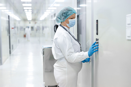 Female scientist in pharmaceutical industry seen standing in front of the door and pushing the button before open it while wearing protective white suit, blue gloves, a face mask and a cap during her night shift in a drug manufacturing.