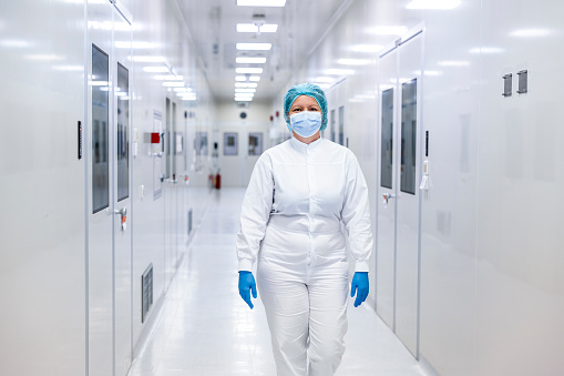 Female employee in pharmaceutical industry seen walking down the hallway and wearing protective white suit, blue gloves, a face mask and a cap during her night shift.