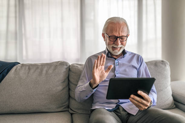 Elderly freelancer video conferencing on digital tablet while sitting at home Elderly freelancer video conferencing on digital tablet while sitting on sofa at home one senior man only stock pictures, royalty-free photos & images