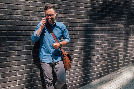 Cheerful male professional wearing formals talking on smart phone while walking against brick wall