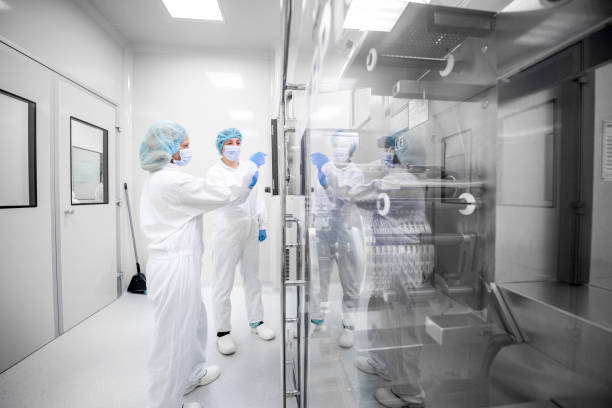 Drug manufacturing - pharmaceutical industry and its female scientists employees Two women wearing safety workwear seen in a pharmaceutical laboratory in front of the complex machine that is the part of drug manufacturing. pharmaceutical manufacturing machine stock pictures, royalty-free photos & images