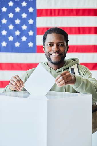 Vertical portrait of smiling black man putting ballot in bin against American flag in background