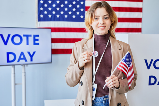 Waist up of smiling young woman holding I vote sticker and American flag while standing in voting station, copy space