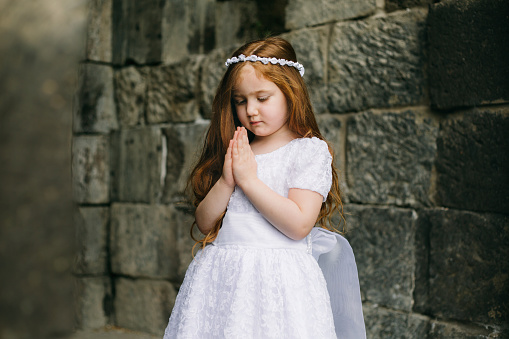 Christianity and spirituality. Adorable redhead girl with white dress praying in front of armenian ancient church. Just baptized girl praying