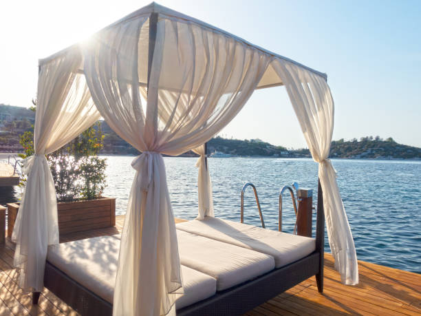 A tranquil beach cabana features flowing white curtains gently swaying in the breeze, framing a spacious seating area adorned with soft cushions. The setting sun casts a warm glow over the ocean or sea, creating a peaceful ambiance. This inviting spot is perfect for relaxation and enjoying the stunning coastal view.