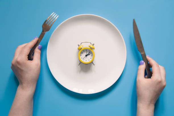 istock Hands holding knife and fork above alarm clock on a plate on blue background. 1405804519