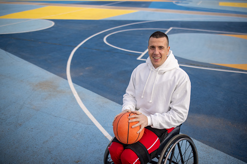 Young disabled Caucasian man in wheelchair, practising the basketball on the basketball outdoor field