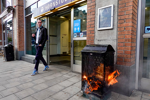 Stockholm, Sweden May 16, 2020 A pedestrian walks by a burning garbage can at the entrance to the Hornstull subway or Tunnelbana station.