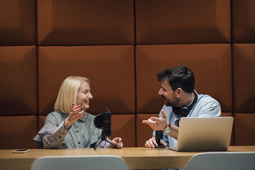 Young radio cohosts at work, doing a radio show, using laptop. Young man and woman talking to a microphone, doing a podcast. Young businesspeople having serious talk while taping it. Interview studio,  anechoic chamber.