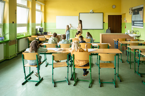 rear view on group of school children in classroom of elementary school, one girl writing on white board beside female teacher