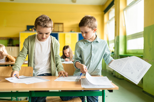 two schoolboys standing at desk and looking together at work sheets on desk in classroom