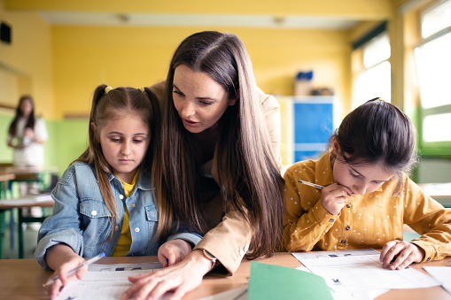 young female teacher standing behind elementary schoolgirls and helping with schoolwork in lesson