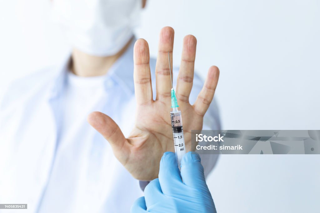 Anti Vaccination Female wearing surgical protective face mask is holding up hand as a stop sign against an injection syringe. Representing anti vaccination movement. Vaccination Stock Photo