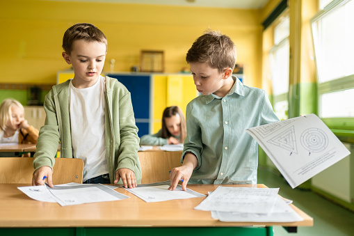 two schoolboys standing at desk and looking together at worksheets on desk in classroom