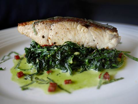 Close up side view of grilled sea bass slice on spinach in white plate, gourmet cuisine menu, dark background