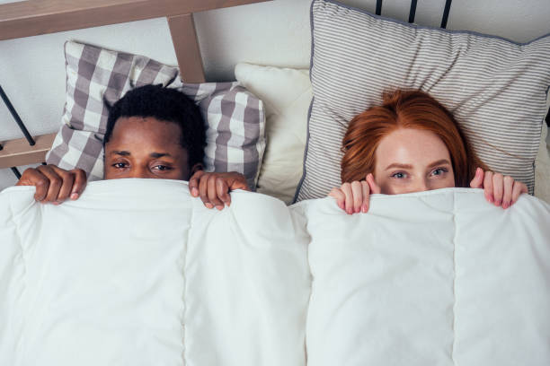 jengibre de pelo rojo caucásico feliz mujer feliz y hombre afro multiétnico juntos acostados en el dormitorio de la cama escondidos bajo la manta.concepto de bashfulness - avergonzamiento fotografías e imágenes de stock