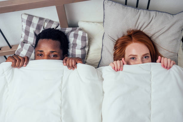 jengibre de pelo rojo caucásico feliz mujer feliz y hombre afro multiétnico juntos acostados en el dormitorio de la cama escondidos bajo la manta.concepto de bashfulness - couple black american culture african culture fotografías e imágenes de stock