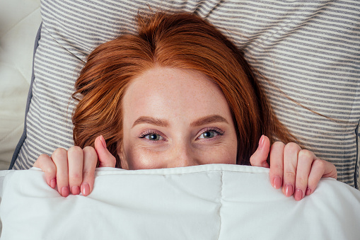 caucasian beautiful redhaired ginger woman hidden under white duvet looking at camera , top view.