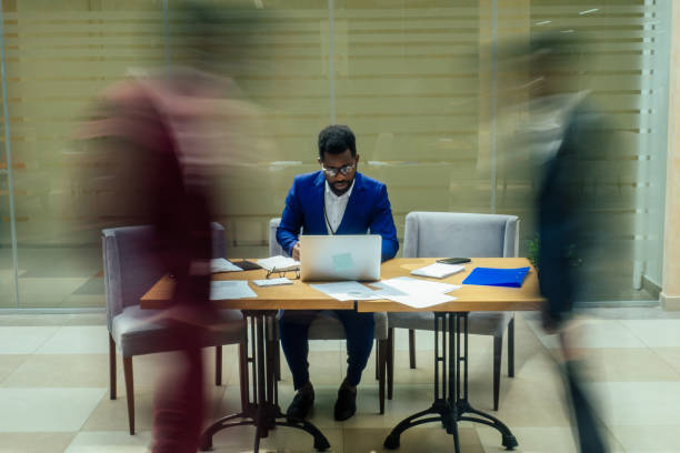 uomo d'affari americano africano che lavora con il computer portatile, persone sfocate che camminano accanto a lui in un grande ufficio - defocused crowd blurred motion business foto e immagini stock