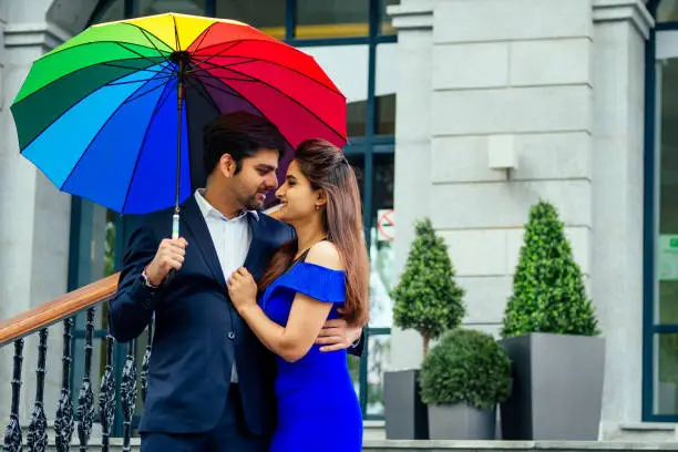 indian woman in long evening blue dress spinding romantic time together with lover handsome boyfriend under multi-colored rainbow umbrella europe urban downtown city.