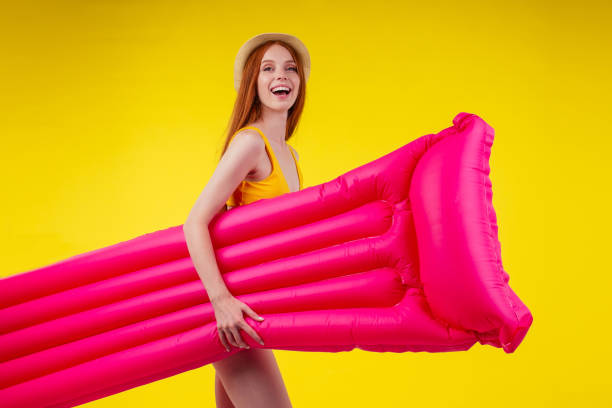 happy redhaired ginger woman ready to pool party,with pink mattress in studio wall yellow background wearing straw hat stock photo