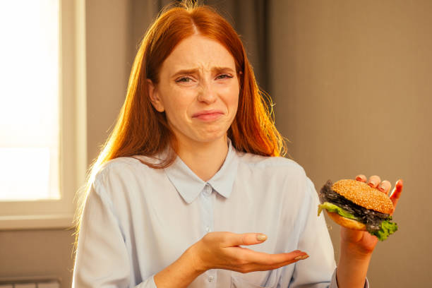 ragazza di zenzero dai capelli rossi che mangia hamburger finto con panino al sesamo e plastica cellophane film spazzatura, acqua potabile da tazza usa e getta con tubo non ecologico nella cucina di casa - white food and drink industry hamburger cheeseburger foto e immagini stock