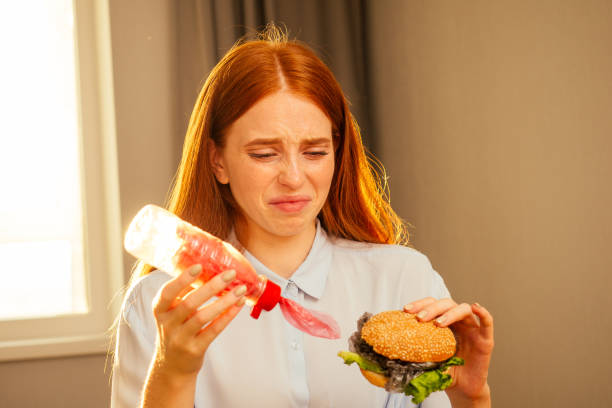 fille au gingembre aux cheveux roux mangeant un faux hamburger avec du pain sésame et des déchets de film de cellophane en plastique, eau potable de la tasse jetable avec tube non écologique à la cuisine à la maison - white food and drink industry hamburger cheeseburger photos et images de collection