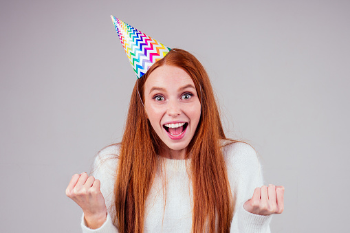 amazed and surprised redhaired ginger woman wearing birthday cap horn holding many gift boxes in studio white background.