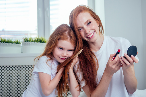 cute foxy hair little girl applying make up and doing hairstyle to redhaired ginger mother at bedroom beautican girlish spa day.