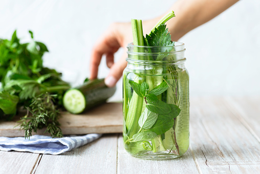 Infused water bottle with an abundance cucumber slices, mint leafs, celery stalk and rosemary in on white wood. Hand of an unrecognizable person in the background is preparing the bottle with more green vegetables.