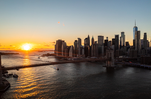 Aerial view of Brooklyn bridge and modern downtown skyscrapers against romantic sunset. Rays of sun reflecting on water surface. Manhattan, New York City, USA.