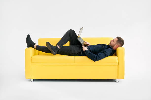 Relaxed man lying on yellow sofa using laptop stock photo