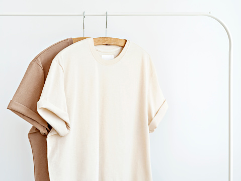 Two t-shirts in neutral milky colours hanging on white rack against white wall. Blank template, mockup for textile design. Summer minimalist outfit idea. Capsule wardrobe.