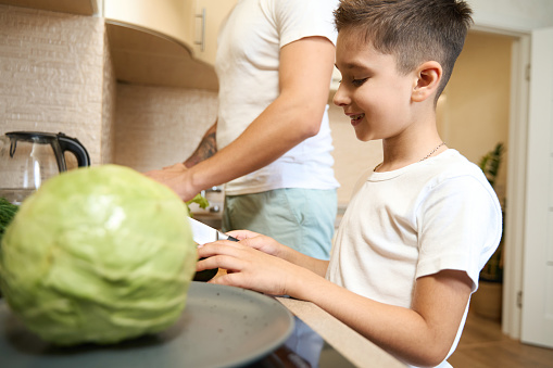 Male kid smiling while cutting vegetables on wooden board in the kitchen near his father