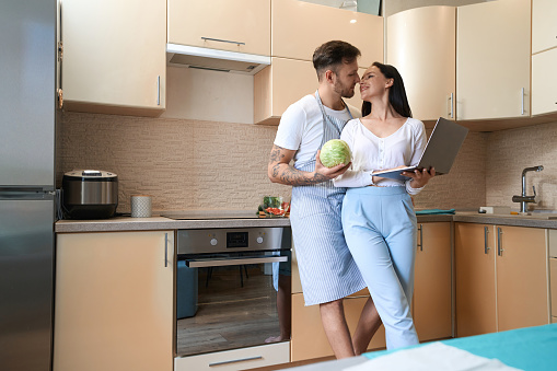 Joyful lady smiling at her boyfriend with cabbage in kitchen while standing beside him with laptop
