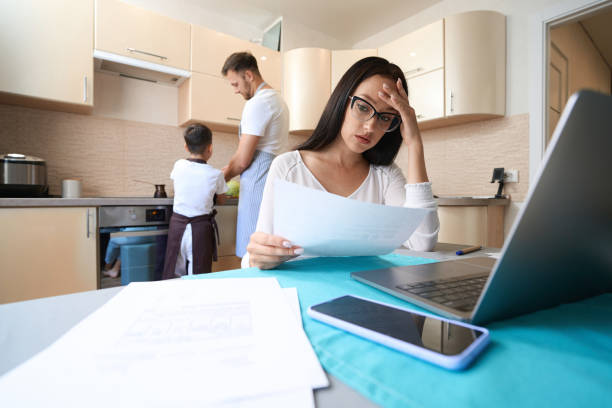istock Tired female checking paper while working on laptop in kitchen 1405862122
