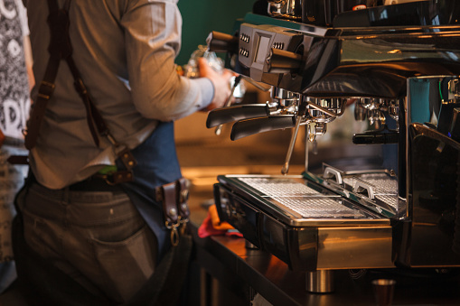 Selective focus shot of a modern, heavy duty espresso machine on a bar counter and unrecognizable barista preparing coffee.