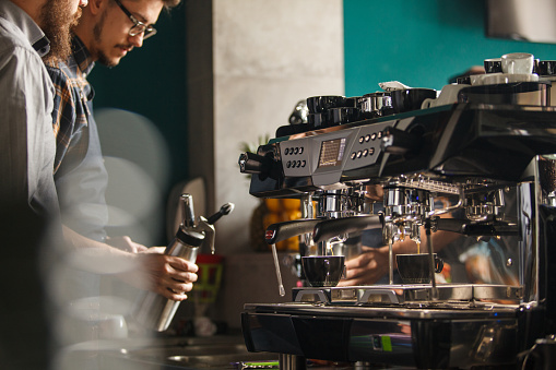 Espresso machine with two cups being poured in focus with two male baristas standing behind bar counter, preparing drinks.