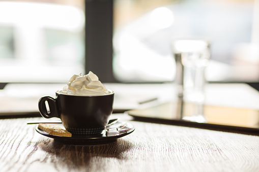 Close up shot of a cup of coffee with whipped cream on top, a bag of sugar and a biscuit, served on a table at the coffee shop.