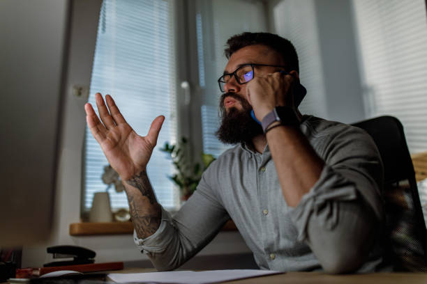 Annoyed businessman gesturing while talking on the phone with a client Low angle view of annoyed businessman sighing and gesturing while talking on the phone with a client when staying late at the office. sighing stock pictures, royalty-free photos & images