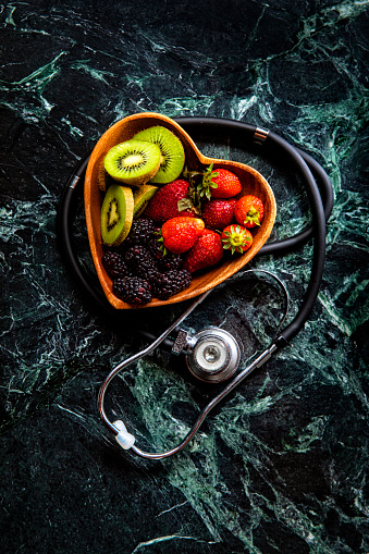 This is an overhead photo of a heart bowl of berries on a green marble background with a doctor stethoscope