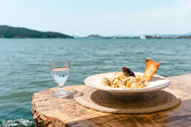 Photo of Seafood pasta spaghetti and a glass of water standing on a wooden table in outside cafe restaurant with scenery sea in the background. Copy blank space. Fish diving into noodles concept