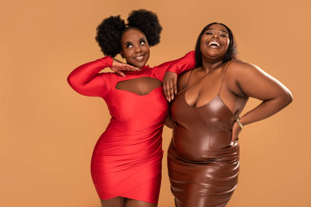 Plus Size african two women posing in party dresses, having fun together, dancing and smiling. Fashionable girls. Body positive and conscious. Plus Size african two women posing in party dresses, having fun together, dancing and smiling.  Studio Background. Real people lifestyle. Fashionable girls. Body positive and conscious. A lot of copy space. body positive couple stock pictures, royalty-free photos & images