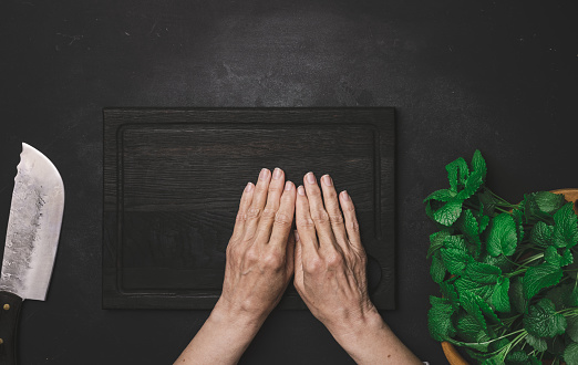 Green fresh mint leaves on a wooden board and two female hands on a black wooden table, top view