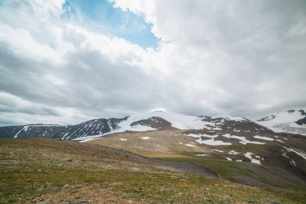 istock Dramatic alpine landscape with large snowy mountain dome in sunlight under clouds in blue sky. Panoramic view to sunlit high snow mountain in dome shape in center. Changeable weather at high altitude. 1405906895