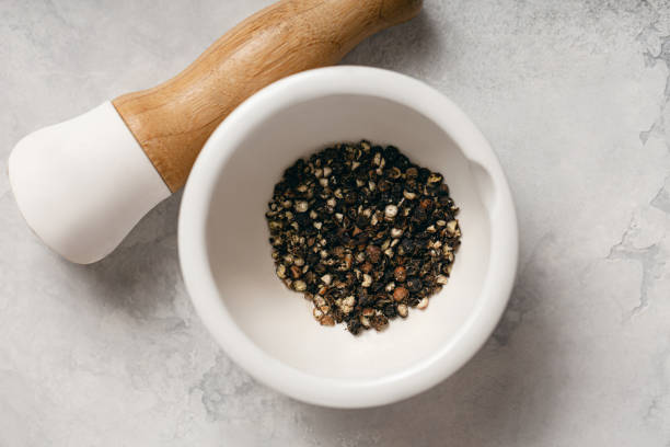 Close-up view of various kinds of pepper in stone mortar. Grinding pepper in mortar with pestle. Several peppercorns falling out from mortar.
