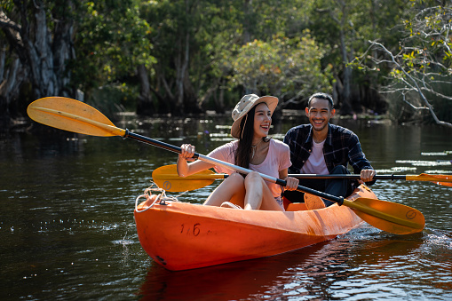 Asian attractive romantic young couple rowing kayak in a forest lake. Backpacker man and woman travel and kayaking on canoe in beautiful mangrove forest enjoy spend time on holiday vacation together.